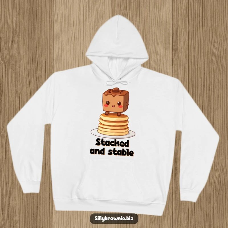 Funny hoodie depicting a brownie character grinning while balancing on a wobbly stack of pancakes, perfect for cozying up.